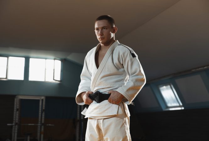 Young judo caucasian fighter in blue kimono with black belt posing confident in the gym, strong and healthy. Practicing martial arts fighting skills. Overcoming, reaching target, self building up.